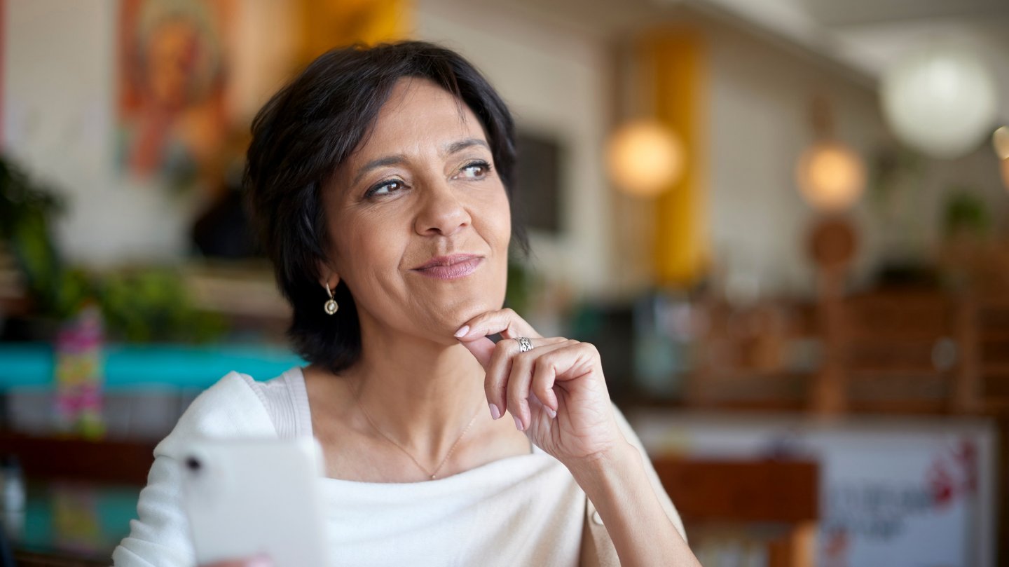  Phone, thinking and happy woman in cafe, restaurant or shop online review, social media or post ideas. Mature, Indian person on smartphone or cellphone with smile for customer service, e commerce app