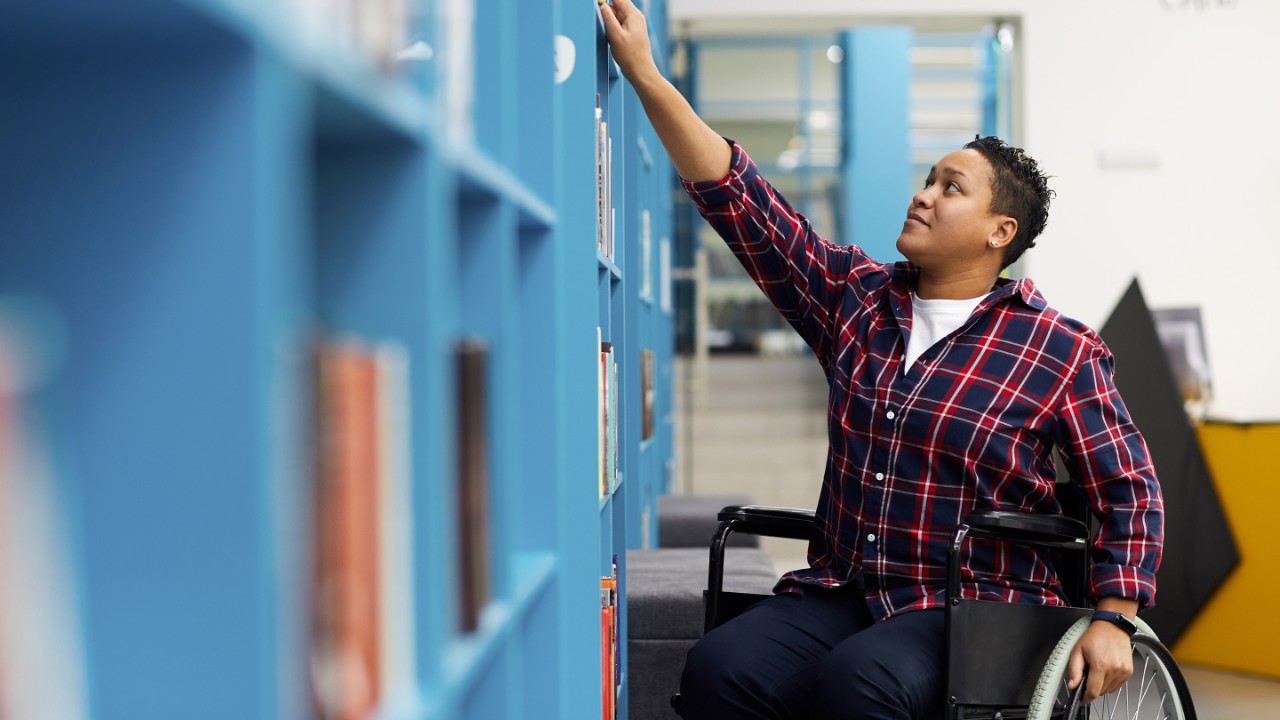 Portrait of disabled student in wheelchair choosing books while studying in college library, copy space