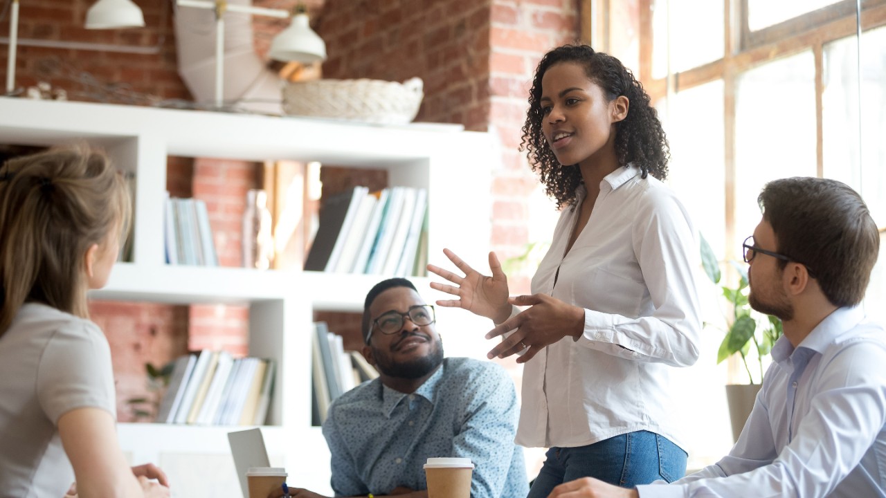 Ambitious smart african black female employee speaking at diverse meeting share creative idea opinion at group briefing while jealous envious skeptical male coworkers looking listening to colleague