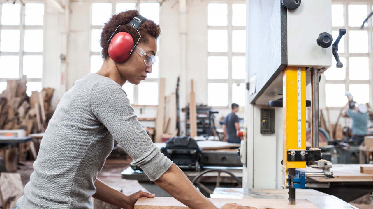 Black woman factory worker using a band saw to cut wood in a woodworking factory.