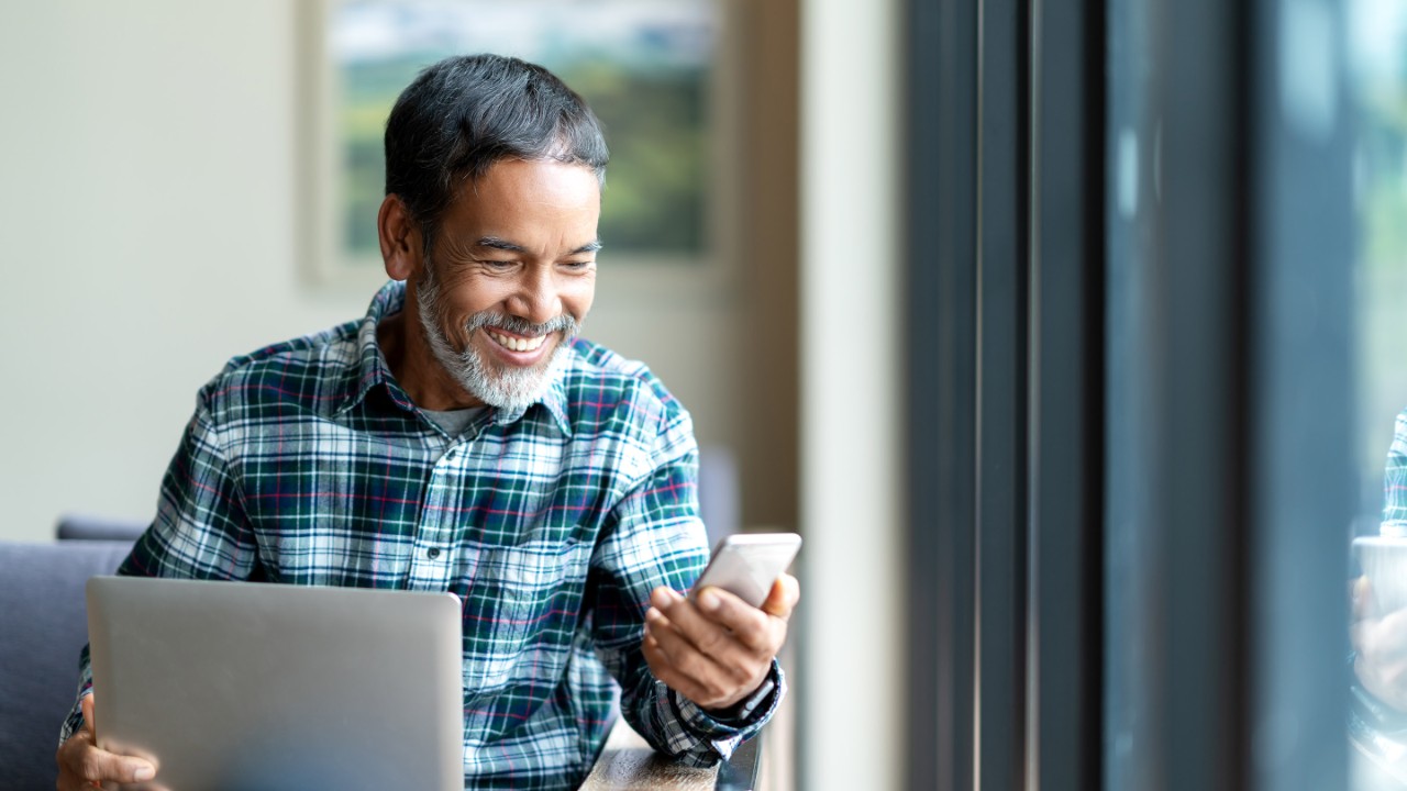 Seated man smiles at phone with laptop on lap