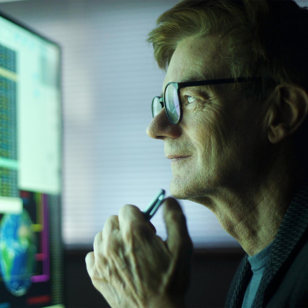 Stock photo of a mature man working in his home office. He’s studying a large computer monitor displaying a variety of numerical data, global information & text.