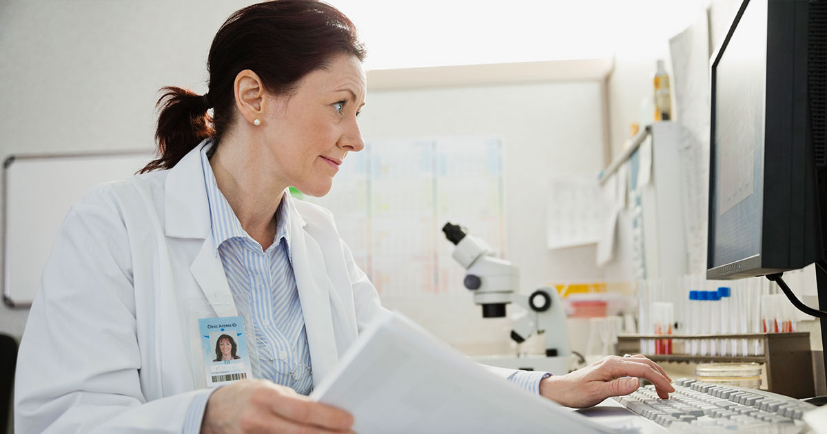 Doctor using computer while reviewing records at desk in clinic, individual, woman