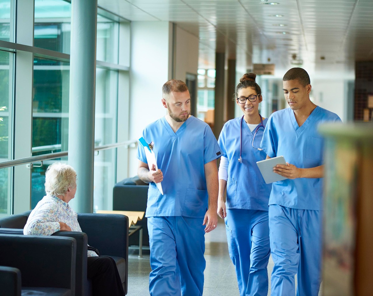 506479358 three junior doctors walking along a hospital corridor discussing case and wearing scrubs. A patient or visitor is sitting in the corridor as they walk past .