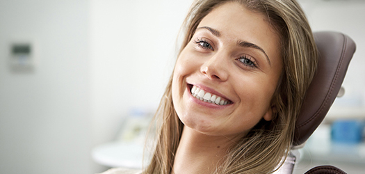 A bright toothy smile and eye contact from a pretty young woman proud to show off her perfect teeth while sitting in a dentist's chair.