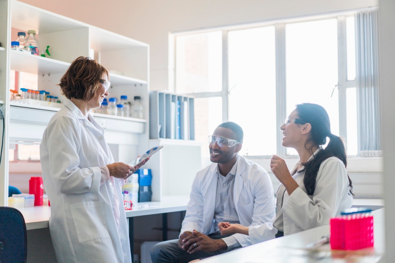 A photo of scientist holding digital tablet while discussing with colleagues. Male and females are wearing protective eyeglasses. They are in brightly lit laboratory.