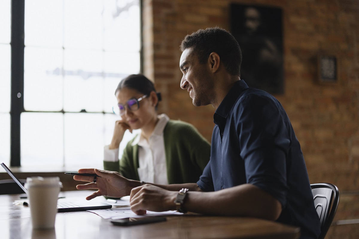 Two young professionals discuss ideas while using a laptop in a modern office. The atmosphere is collaborative, with one person engaged in conversation and the other listening intently.