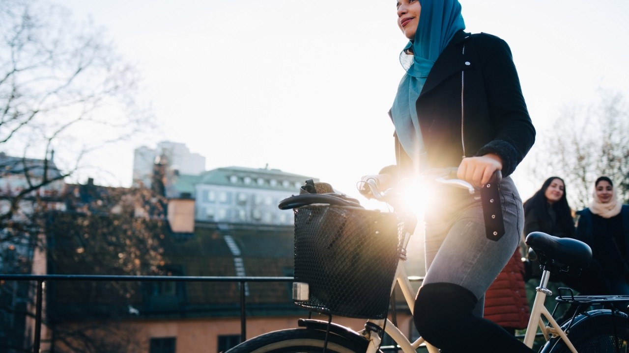 Back lit of young woman cycling by female friends against sky in city