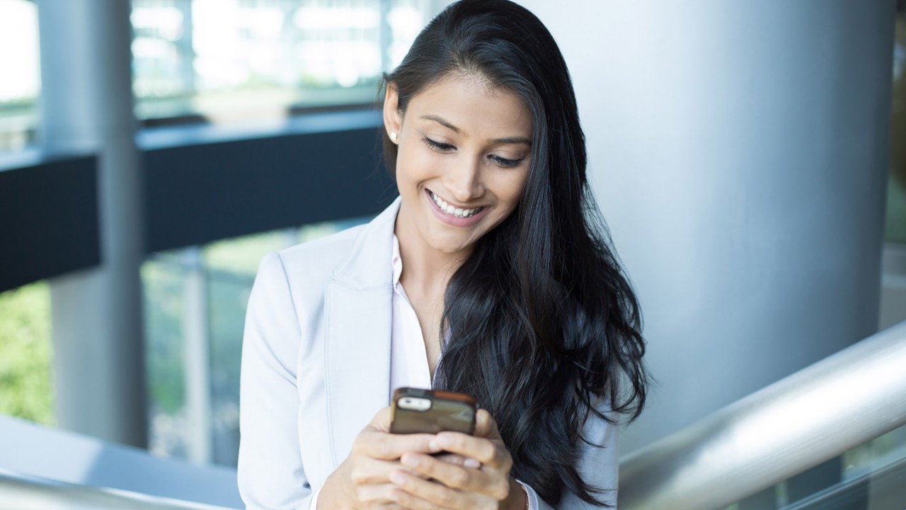 Closeup portrait, young successful happy business woman in light white gray suit, checking her cellphone, isolated on interior indoors office background . Business communication