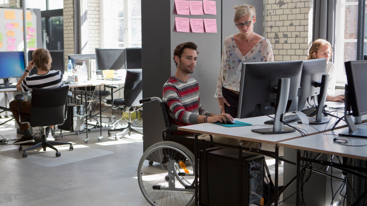 Entorno de oficina con personas trabajando con ordenadores, incluida una persona en silla de ruedas