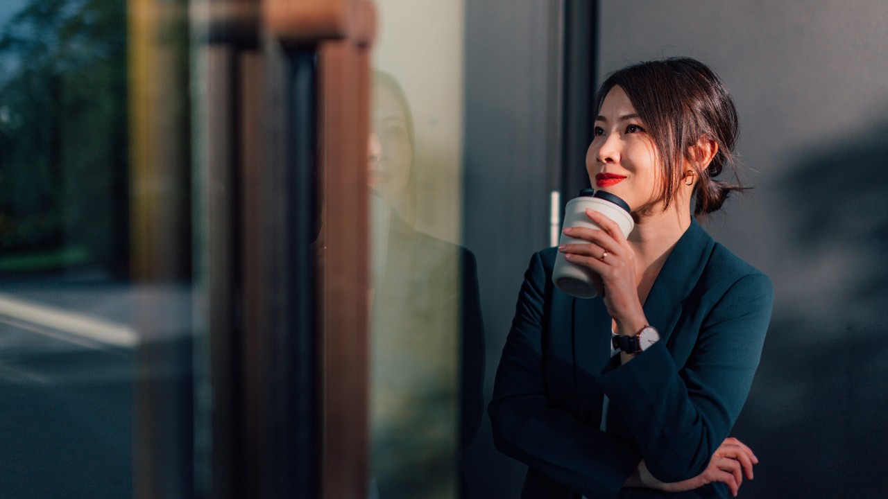 Confident Asian woman looking out the window, holding a reusable coffee cup, determined to succeed.