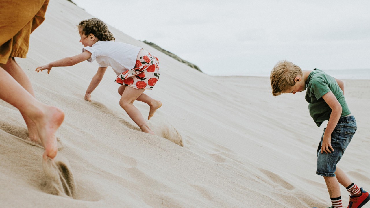Three children clamber up a steep sand dune, which takes a lot of effort as the soft sand gives way under their little feet and hands