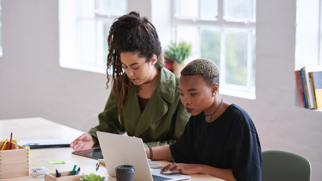 College students working together two young women friends brainstorming ideas for project sitting at desk using laptop computer in class