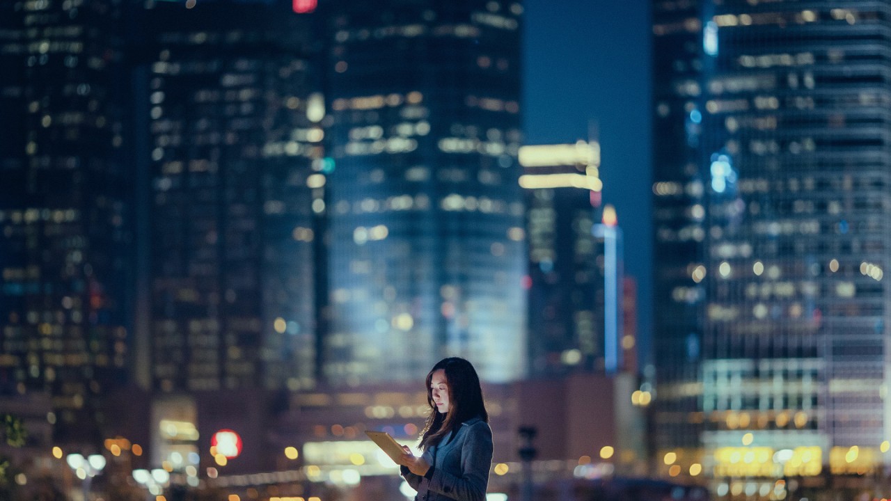Pretty office lady is using digital tablet, standing against illuminated highrise corporate buildings at night time.