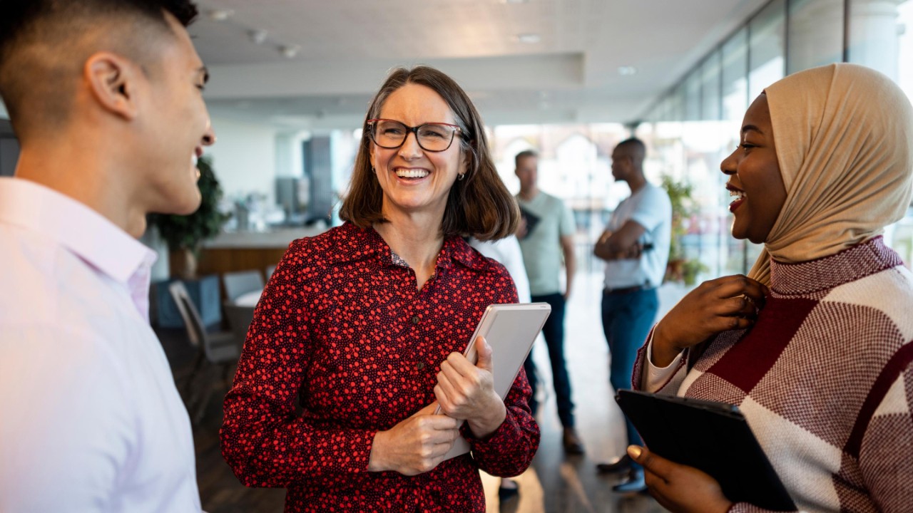 Medium shot of a mixed ethnic group of business professionals at a networking conference/meeting room in the North East of England. They are standing holding books/digital tablets smiling while discussing business together.