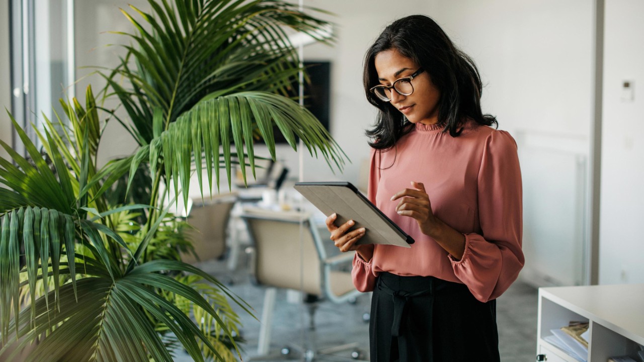 Cadre de sexe féminin aux cheveux noirs de longueur moyenne portant des lunettes et des vêtements de travail devant une salle de réunion moderne et vérifiant les données sur une tablette.