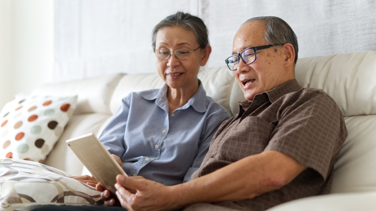 Senior couple sitting on sofa in home playing tablet and relaxing together. They are smiling and enjoy to spend their time together with happiness. Happy retirement life concept.