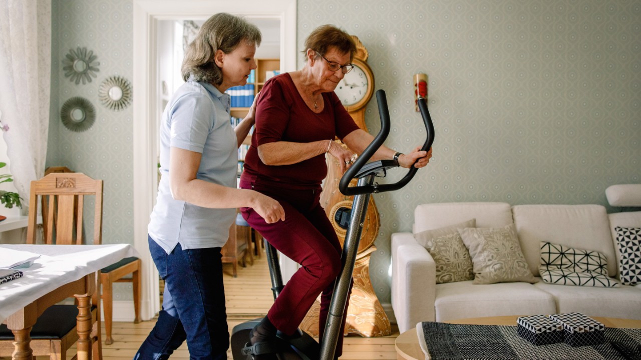 Mature female physical therapist assisting elderly woman on exercise bike at nursing home