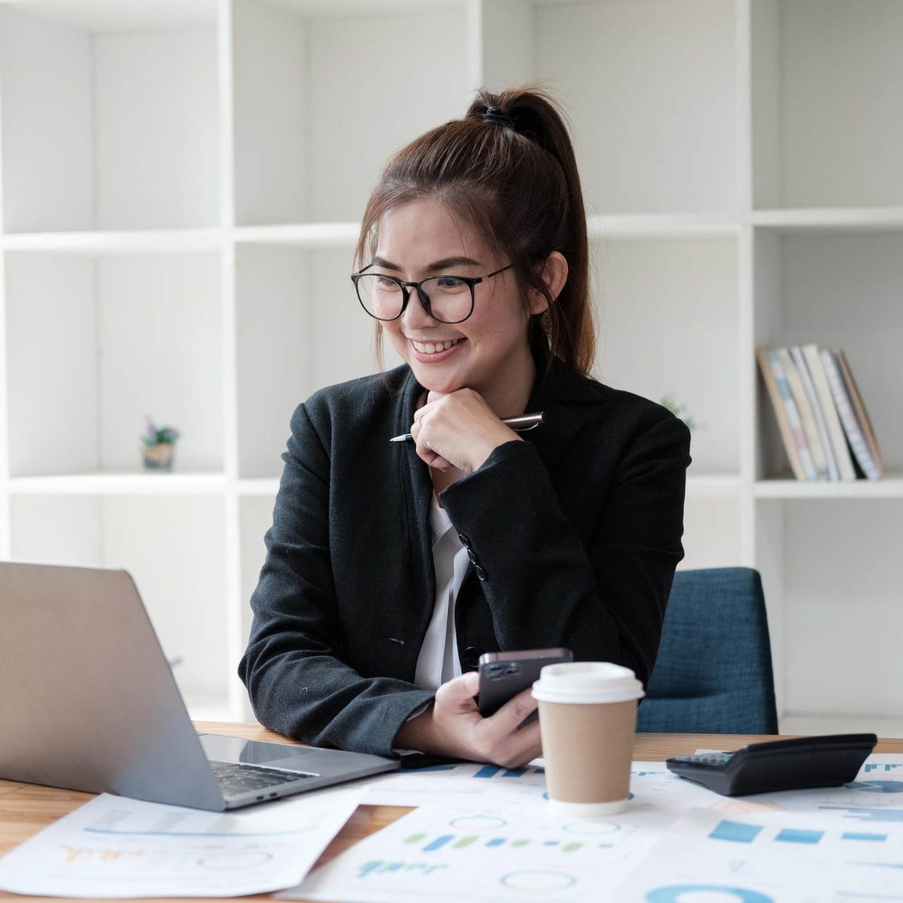 Smart Asian businesswoman working with laptop computer while holding smartphone in her hands, sitting at office desk.