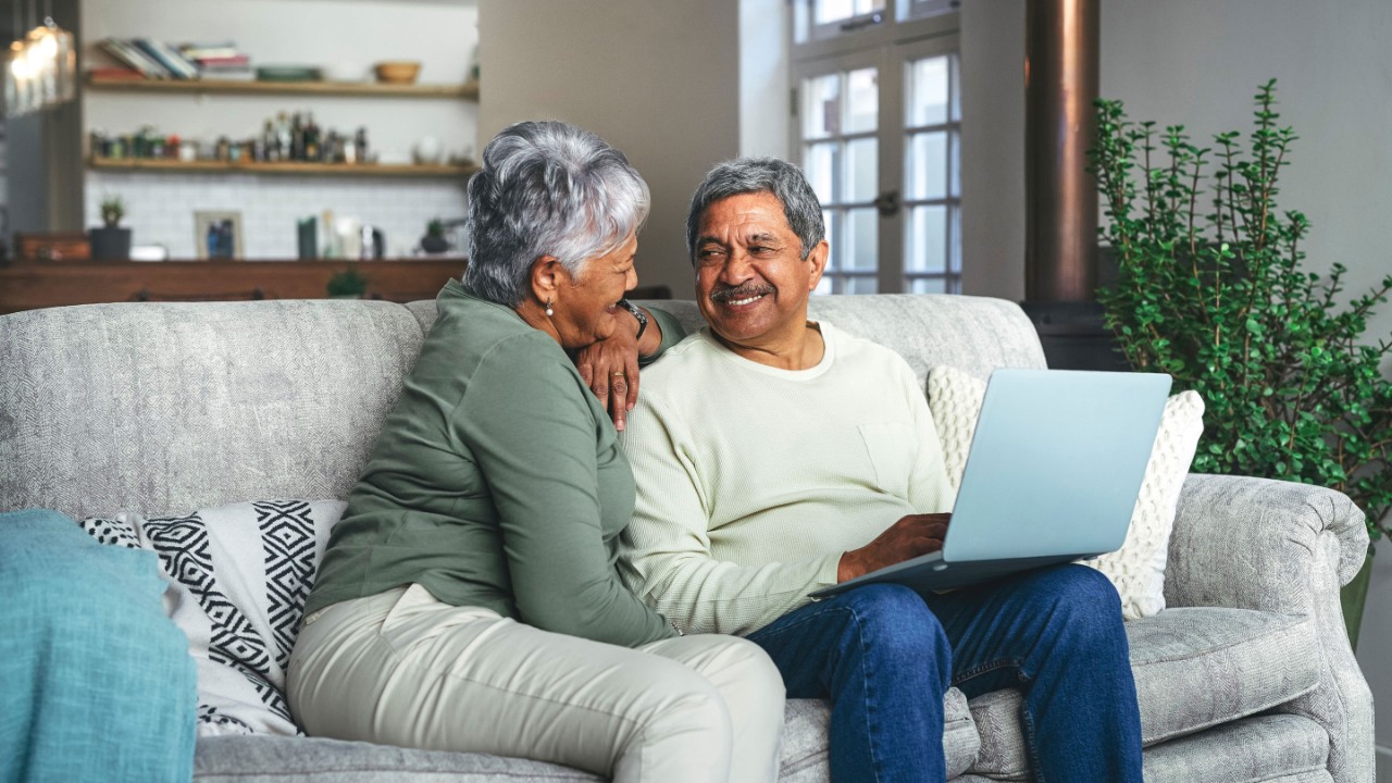 Senior couple smiling each other with laptop at seating on a sofa at home