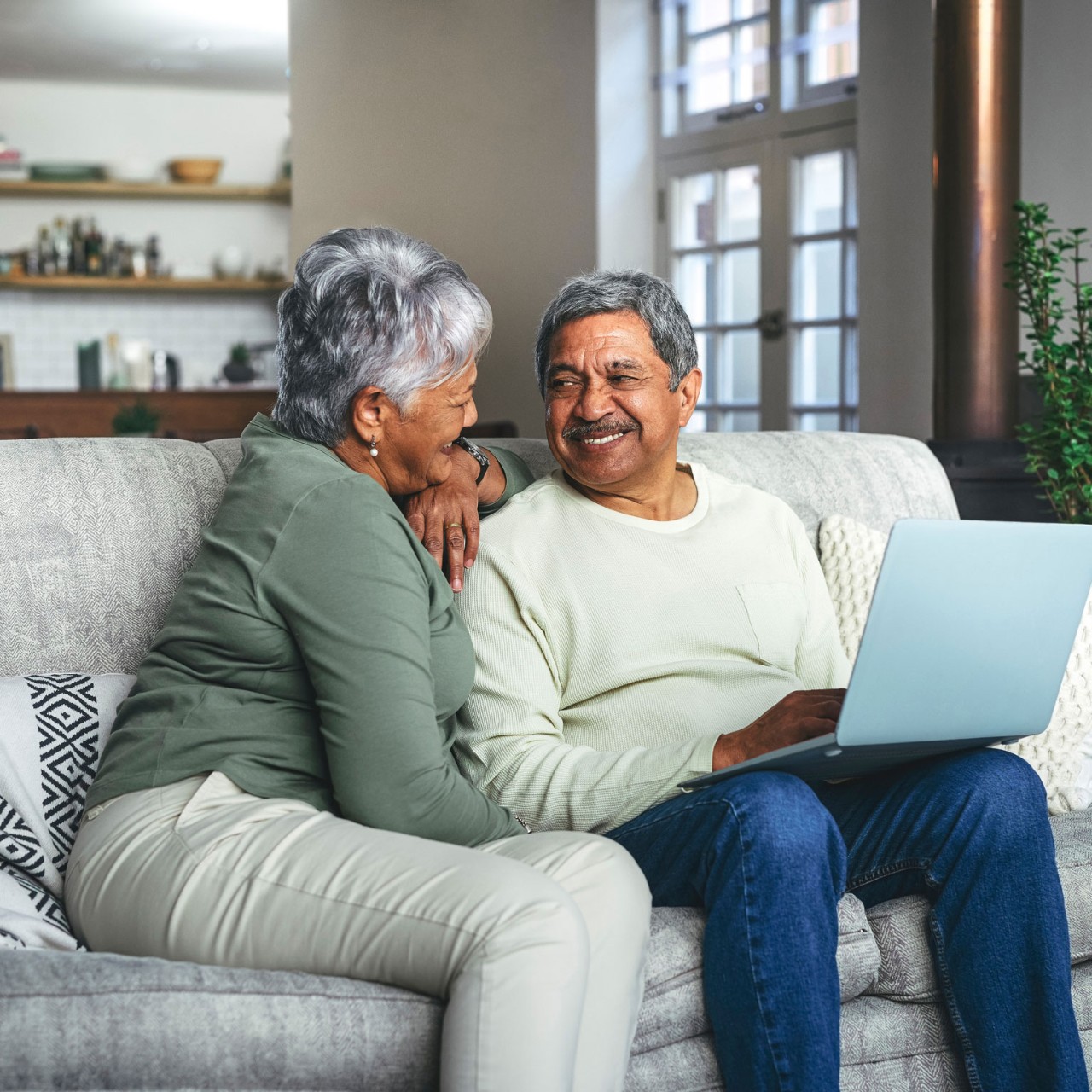 Senior couple smiling each other with laptop at seating on a sofa at home