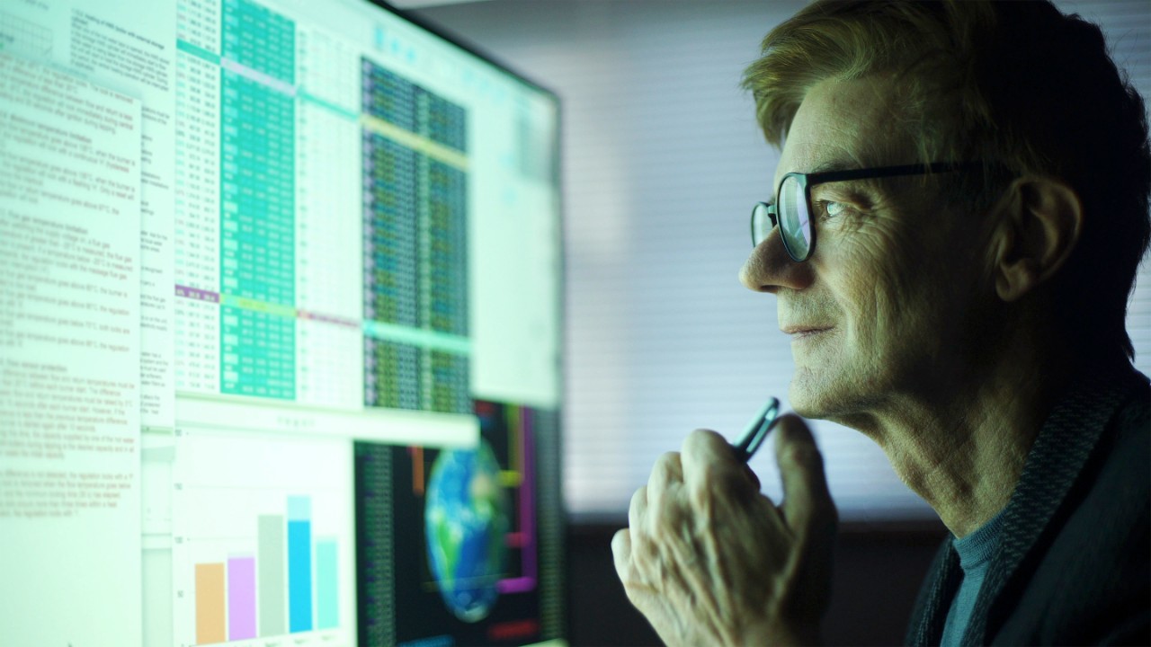 Stock photo of a mature man working in his home office. Heâ  s studying a large computer monitor displaying a variety of numerical data, global information & text.