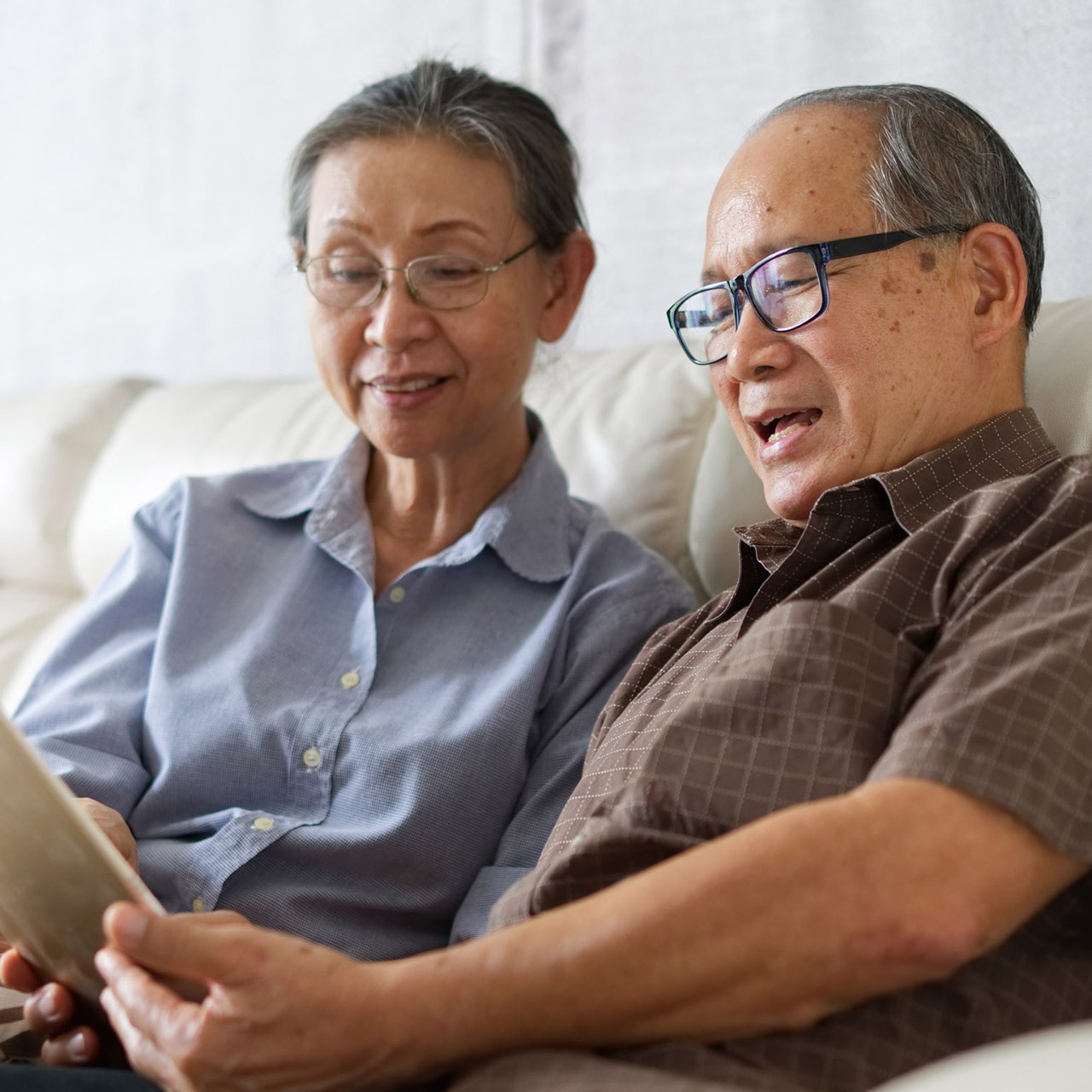 Senior couple sitting on sofa in home playing tablet and relaxing together. They are smiling and enjoy to spend their time together with happiness. Happy retirement life concept.