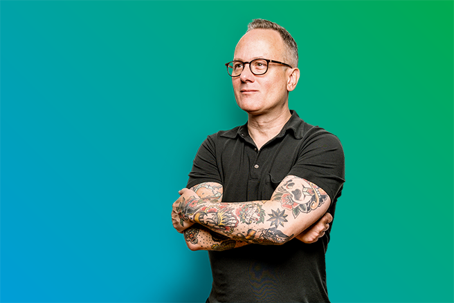Male standing with folded arms covered in tattoos, wearing glasses - blue to green gradient