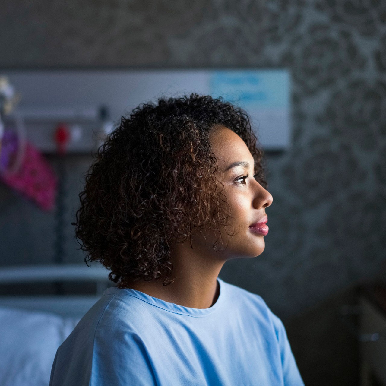 Young woman with curly hair sitting at hospital. Female patient is on bed in ward. She is wearing blue gown.