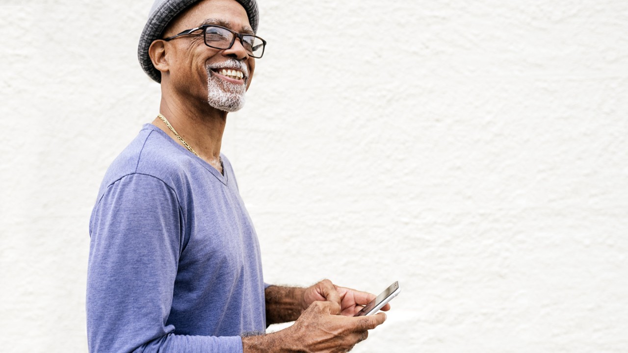 Stock photo of mature afro man standing in front of a white wall. He is looking away from the camera. He is wearing glasses and a hat.