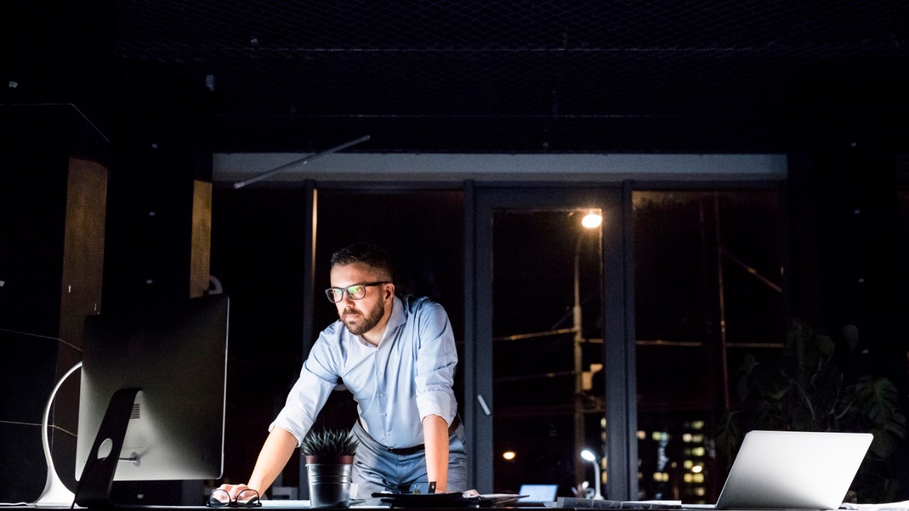 Thoughtful hipster businessman in his office working late at night at desk with computer and laptop.