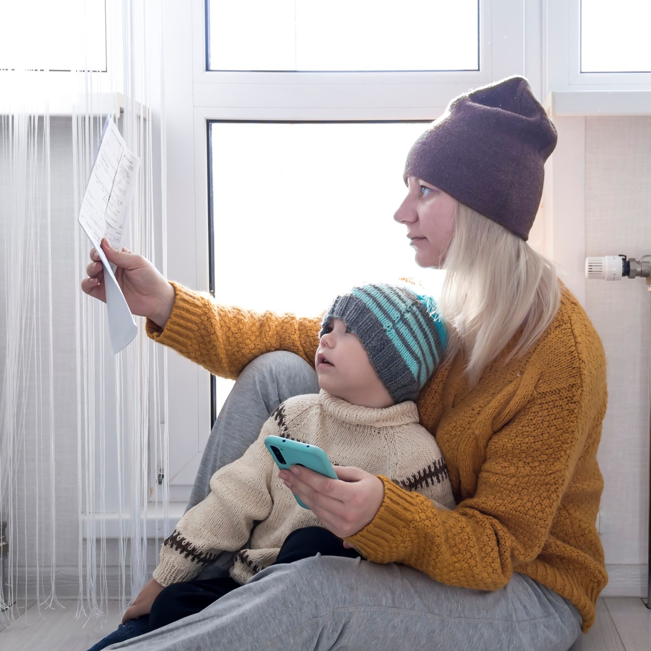 .A mother with a small child in a yellow sweater and hats is counting money and thinking how to pay bills and taxes, near a heater with a thermostat.