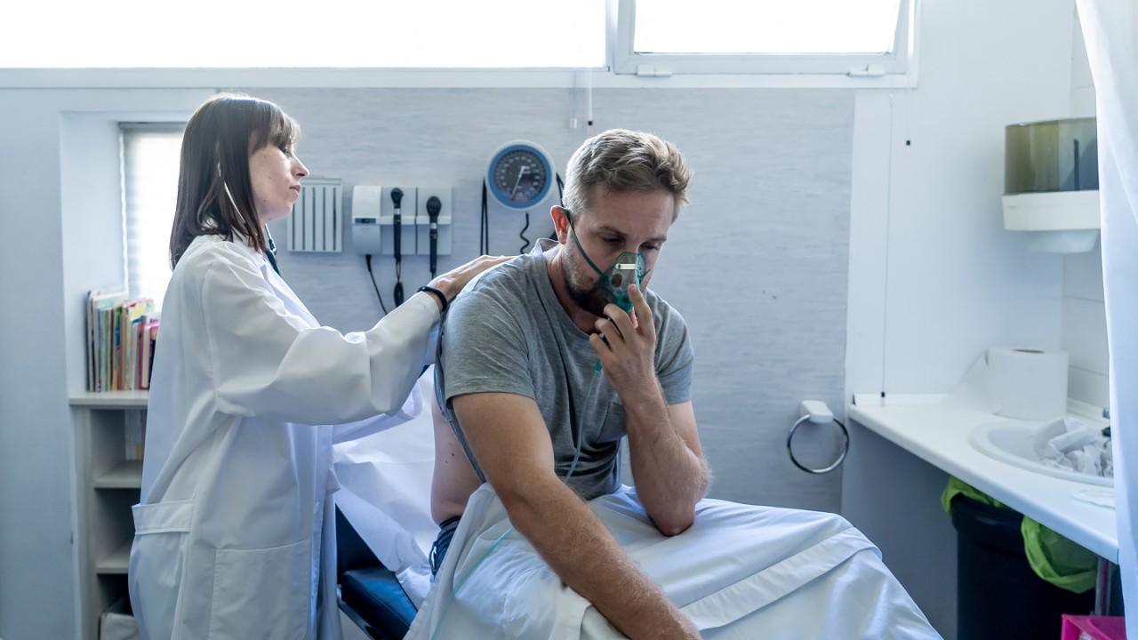 Young sick man patient with Oxygen Mask while female doctor listens his chest with stethoscope in hospital emergency room. In Smoking and respiratory diseases and anti tobacco advertising campaign.
