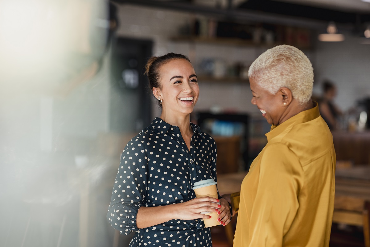 Two women colleagues laughing while standing in a cafe at their workplace. One of the women is holding a take out hot drink cup.
