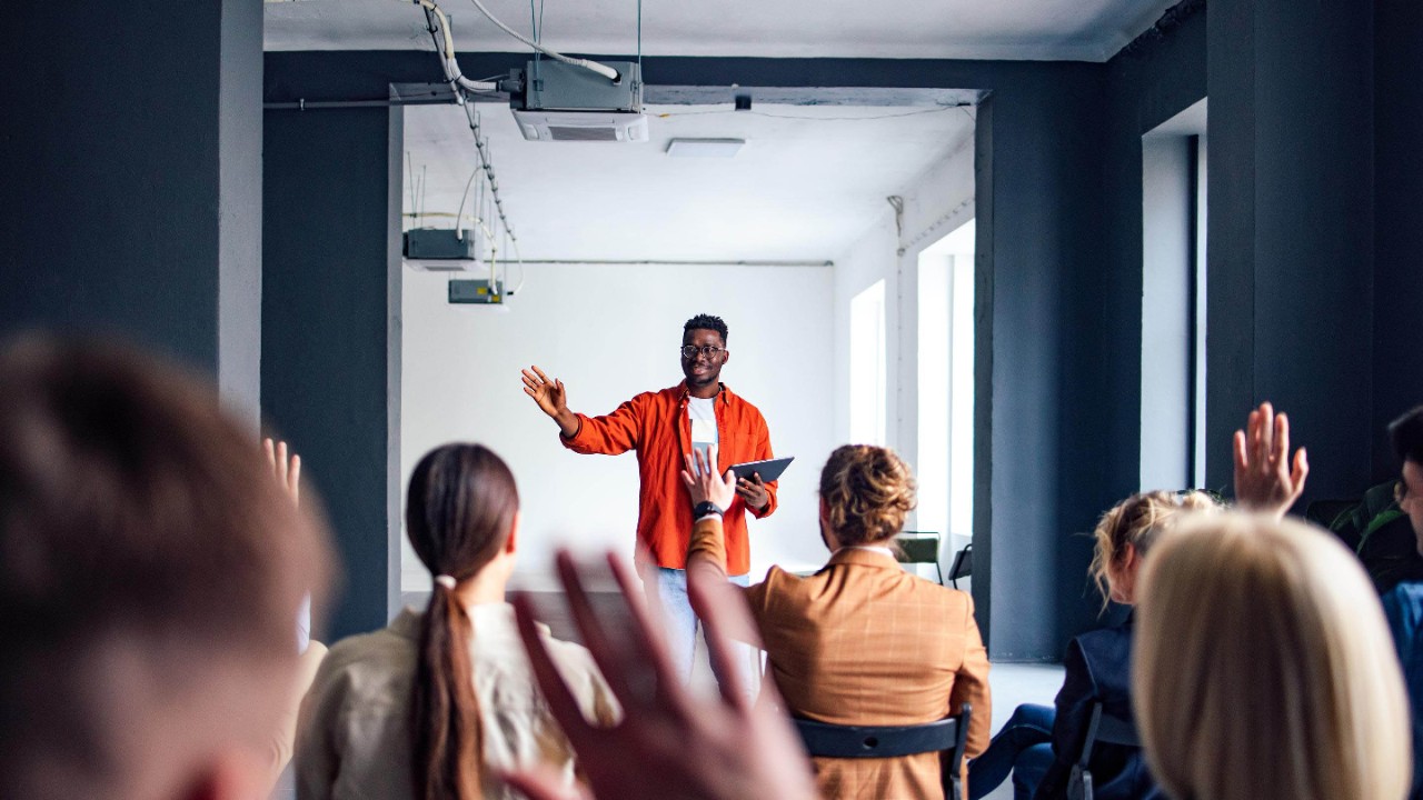 Handsome cheerful man in a orange shirt standing in front of an audience holding a tablet and using hand gestures to interact with the audience.