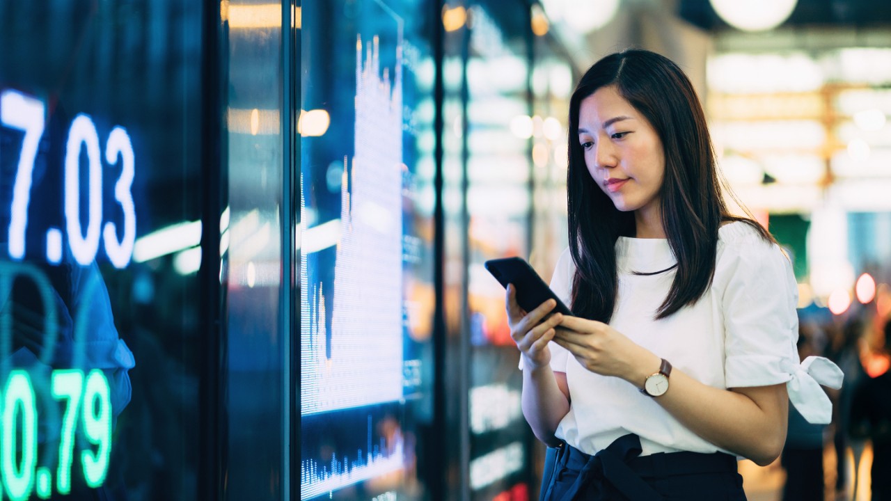 Confidence young Asian businesswoman checking financial trading data on smartphone by the stock exchange market display screen board in downtown financial district