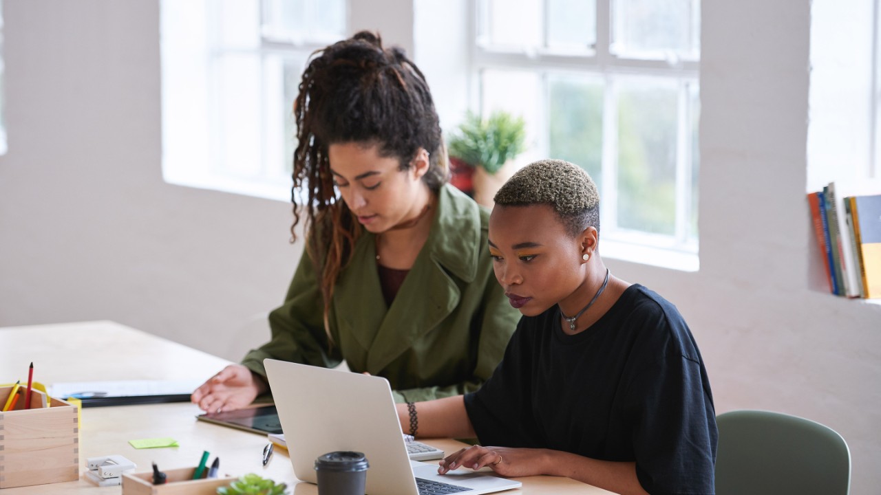 College students working together two young women friends brainstorming ideas for project sitting at desk using laptop computer in class