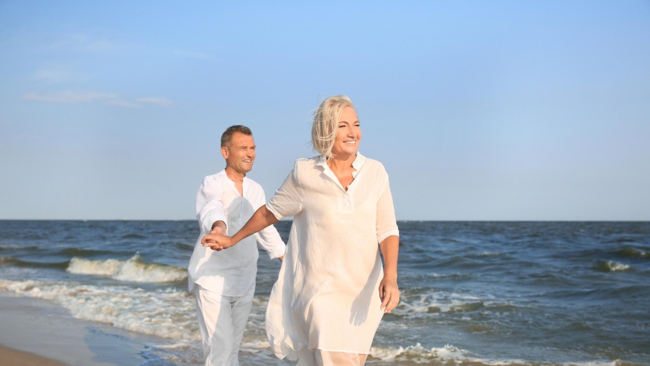 En eldre mann og kvinne kledd i hvitt holder hender på en strand