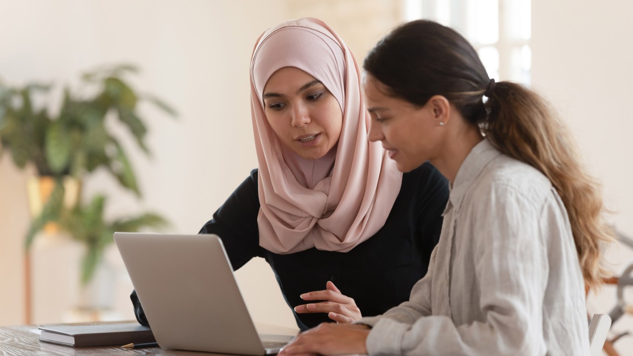 Concentrated young arabian woman in hijab sitting with smiling colleague at table, looking at computer screen, explaining new company software. Focused team leader training millennial female intern.
