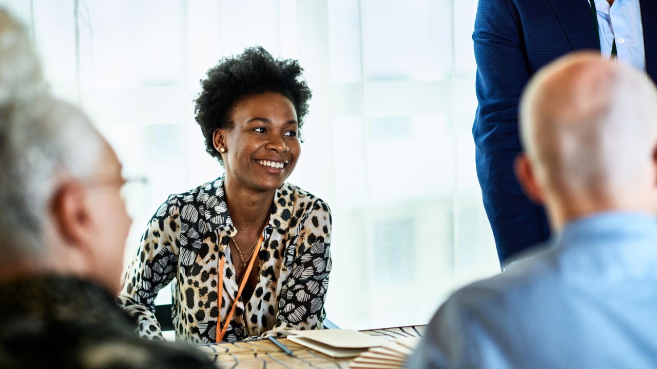 Cheerful woman in her 30s in business meeting with colleagues, smiling and listening, confidence, expertise, team player.