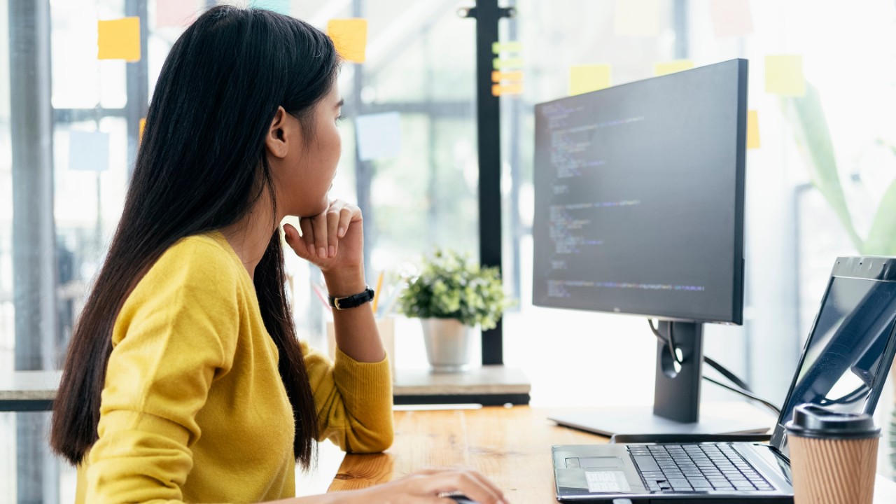 Mulher sentada numa secretária a trabalhar num computador junto a uma grande janela.