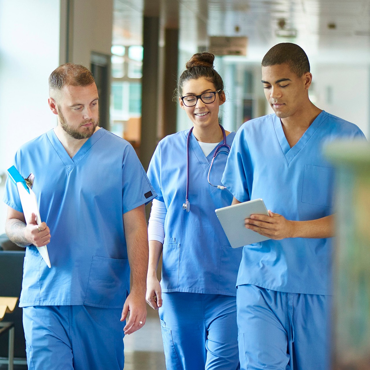 three junior doctors walking along a hospital corridor discussing case and wearing scrubs. A patient or visitor is sitting in the corridor as they walk past .