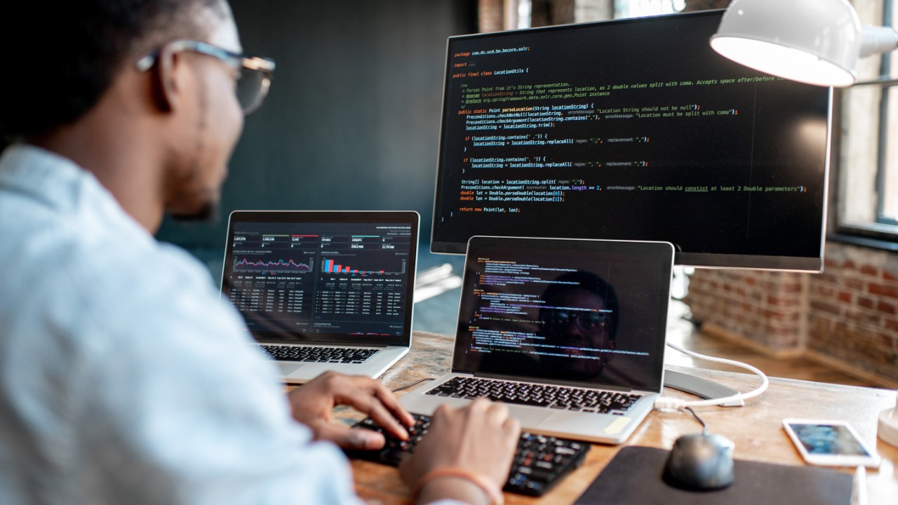 234092554 Young african male programmer writing program code sitting at the workplace with three monitors in the office. Image focused on the screen