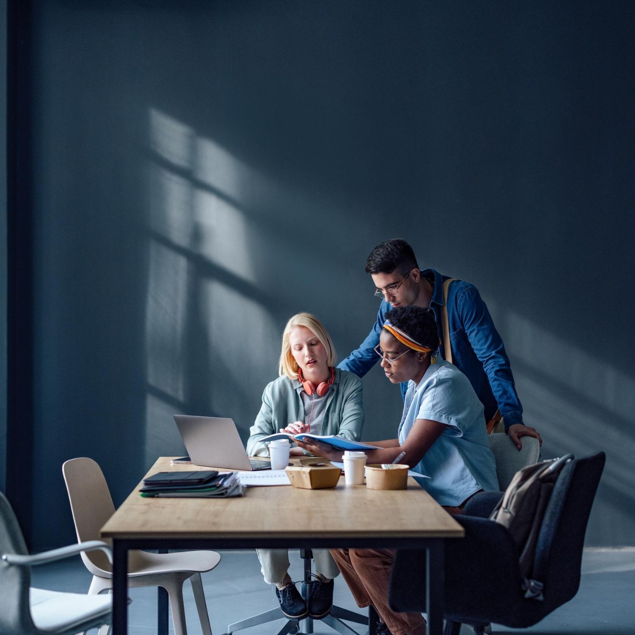 Vários grupos étnicos de amigos que trabalham juntos num projeto escolar enquanto estão sentados e de pé à secretária e leem uma nova lição a partir de livros e computadores portáteis.