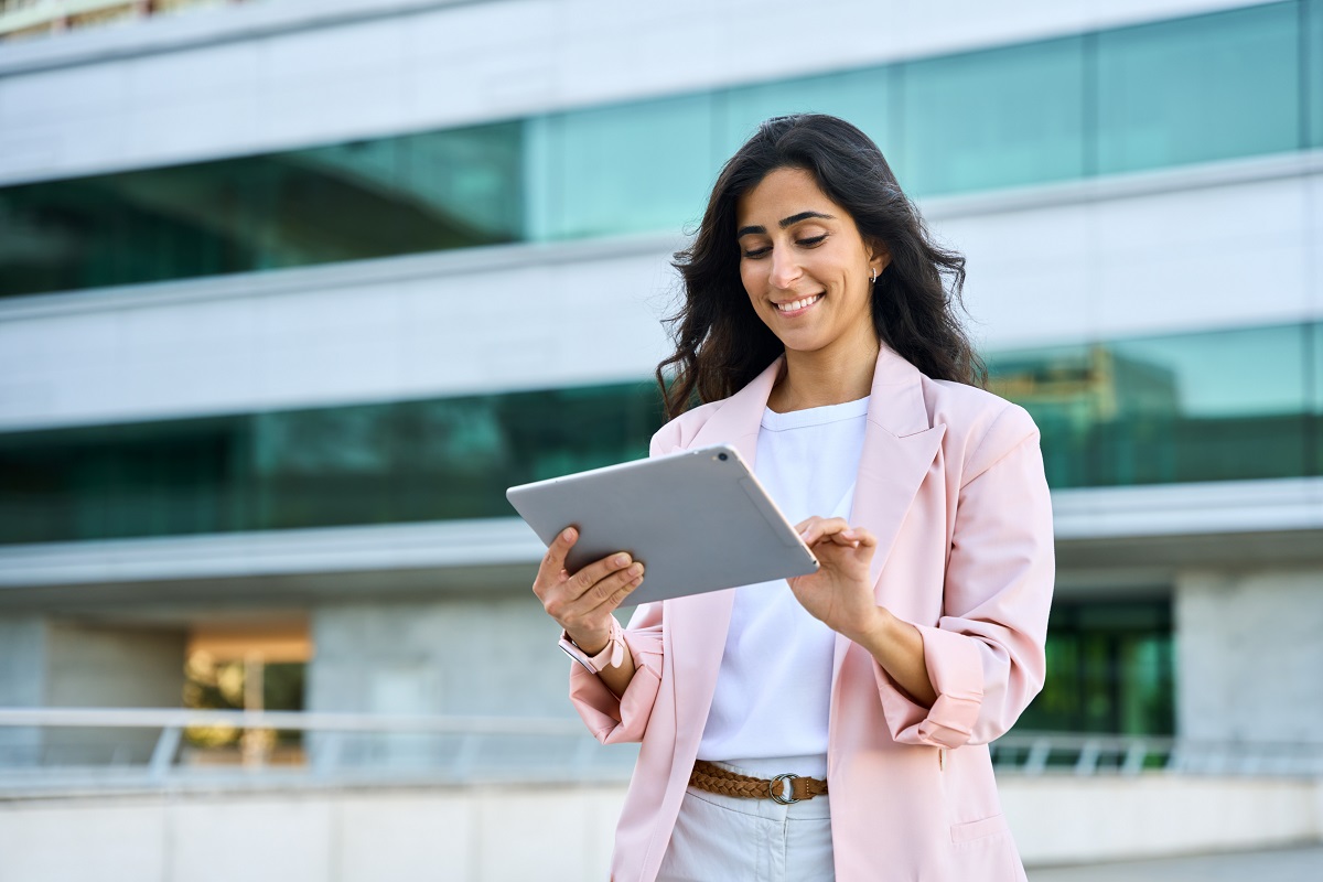 Smiling young middle eastern Israel businesswoman using tablet pc application for online remote work at office business building outdoors. Indian or arabic woman holding digital computer. Copy space