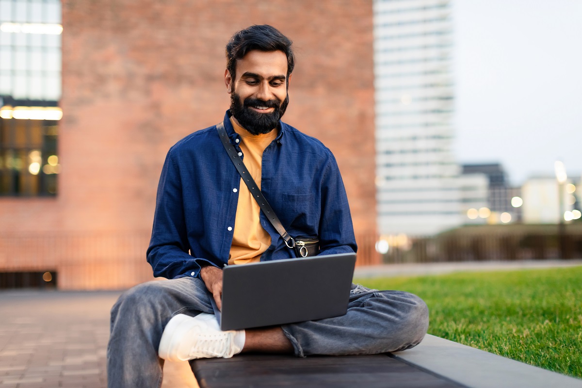 Middle Eastern Guy Freelancer Working Online On Laptop Sitting Outside In Urban Area, Websurfing And Using Website, Advertising Internet Offer. Freelance Career And Technology Concept