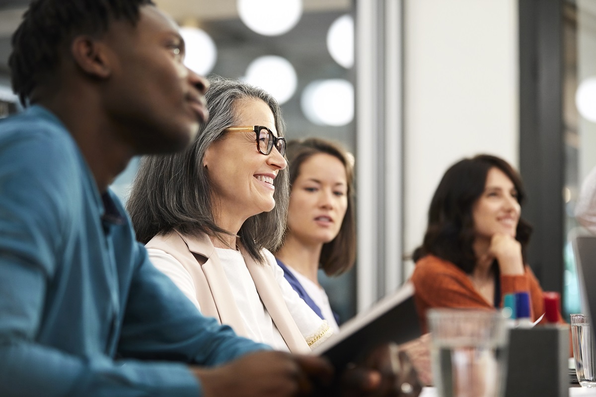 Smiling mature businesswoman sitting with colleagues while looking away. Male and female executives are in board room during meeting. They are at creative office.