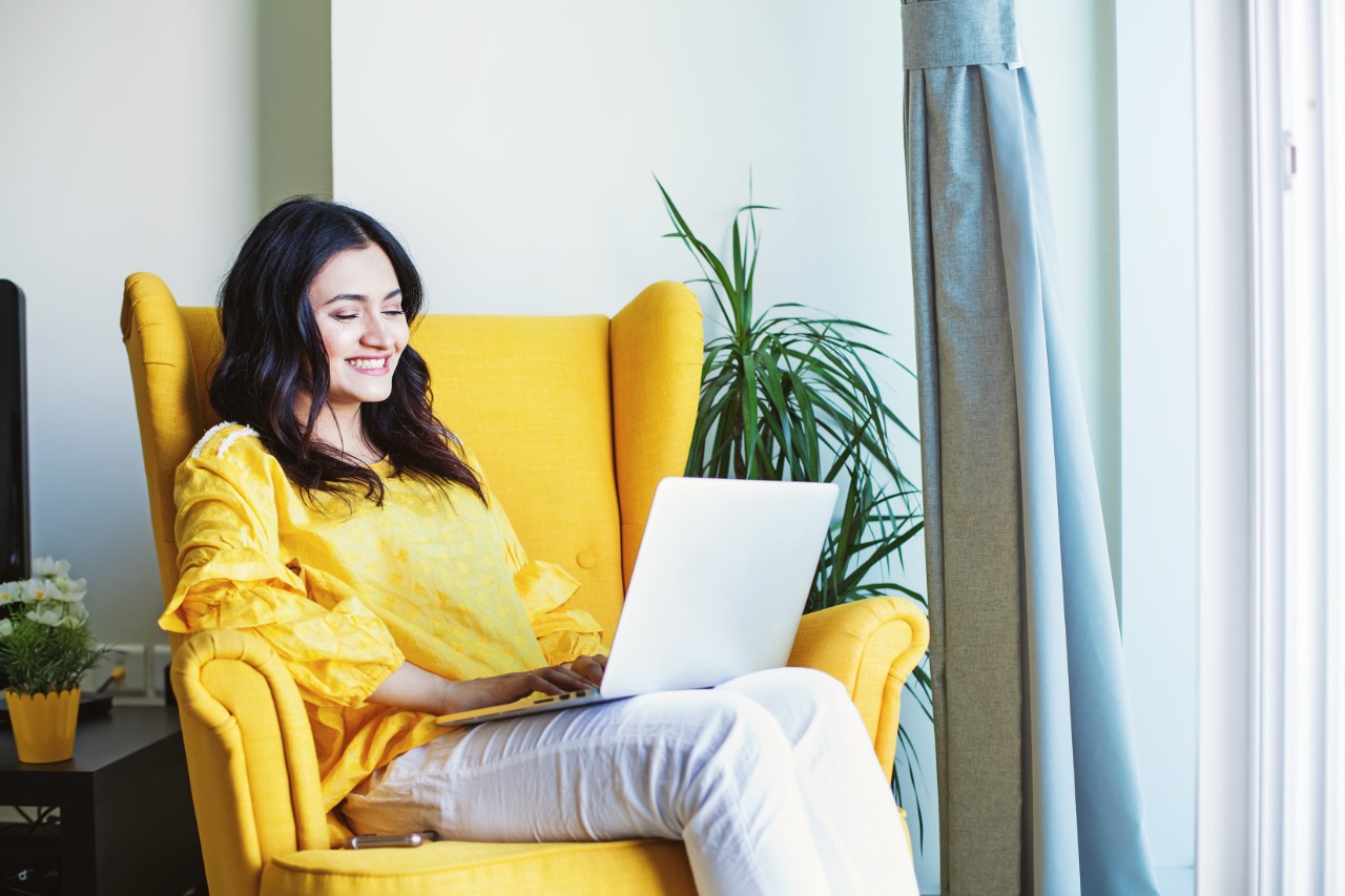 Young Indian woman using her laptop at home while sitting on a yellow armchair